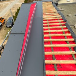 Daniel Komplet Service - Montaż nowej, grafitowej blachy na dachu z widoczną czerwoną membraną i drewnianą konstrukcją łatowania. Widok z góry na dach w trakcie prac dekarskich.