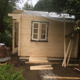 Daniel Komplet Service - Budowa drewnianego domku letniskowego: widoczne ściany z oknem, konstrukcja dachu pod folią ochronną, deski na ziemi, otoczenie zieleni