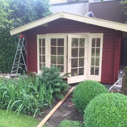 Daniel Komplet Service - Czerwony domek narzędziowy z kremowymi drzwiami i oknami w ogrodzie, drabina oparta o ścianę, osoba pracująca na dachu, widoczne rośliny i drzewo.