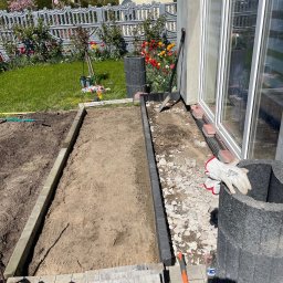 Daniel Komplet Service - Przygotowanie podłoża pod taras: wykopane koryto z betonową obwódką, piasek, gruz, narzędzia i rękawice robocze przy ścianie budynku z dużymi oknami.