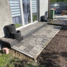 Daniel Komplet Service - Wykończony taras z szarej kostki brukowej przed wejściem do domu, z dekoracyjnymi donicami z betonu architektonicznego i widocznym fragmentem ogrodu.