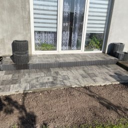 Daniel Komplet Service - Ułożony z szarych kostek brukowych taras przed wejściem do domu z białymi drzwiami balkonowymi, obok ustawione czarne betonowe donice, widoczna świeżo przygotowana ziemia pod trawnik.