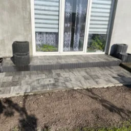 Ułożony z szarych kostek brukowych taras przed wejściem do domu z białymi drzwiami balkonowymi, obok ustawione czarne betonowe donice, widoczna świeżo przygotowana ziemia pod trawnik.