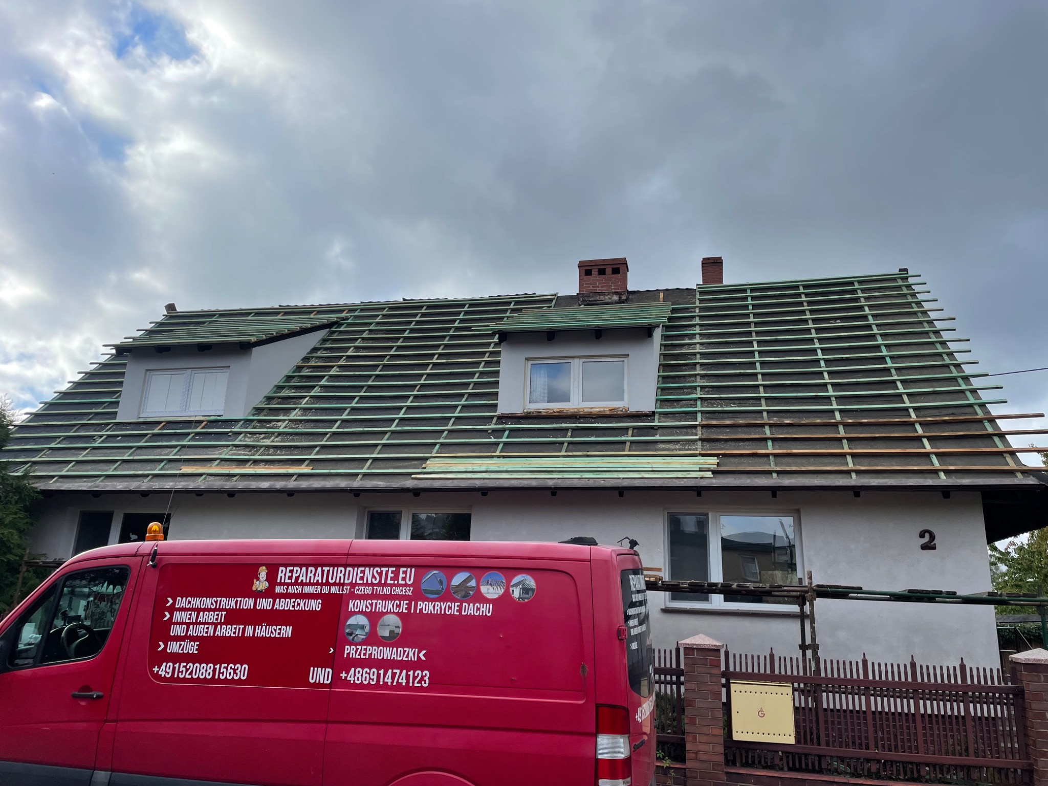 Dom jednorodzinny z częściowo zdjętym starym pokryciem dachu i widoczną konstrukcją nośną z łat, czerwony bus firmy remontowej z reklamą zaparkowany przed posesją.