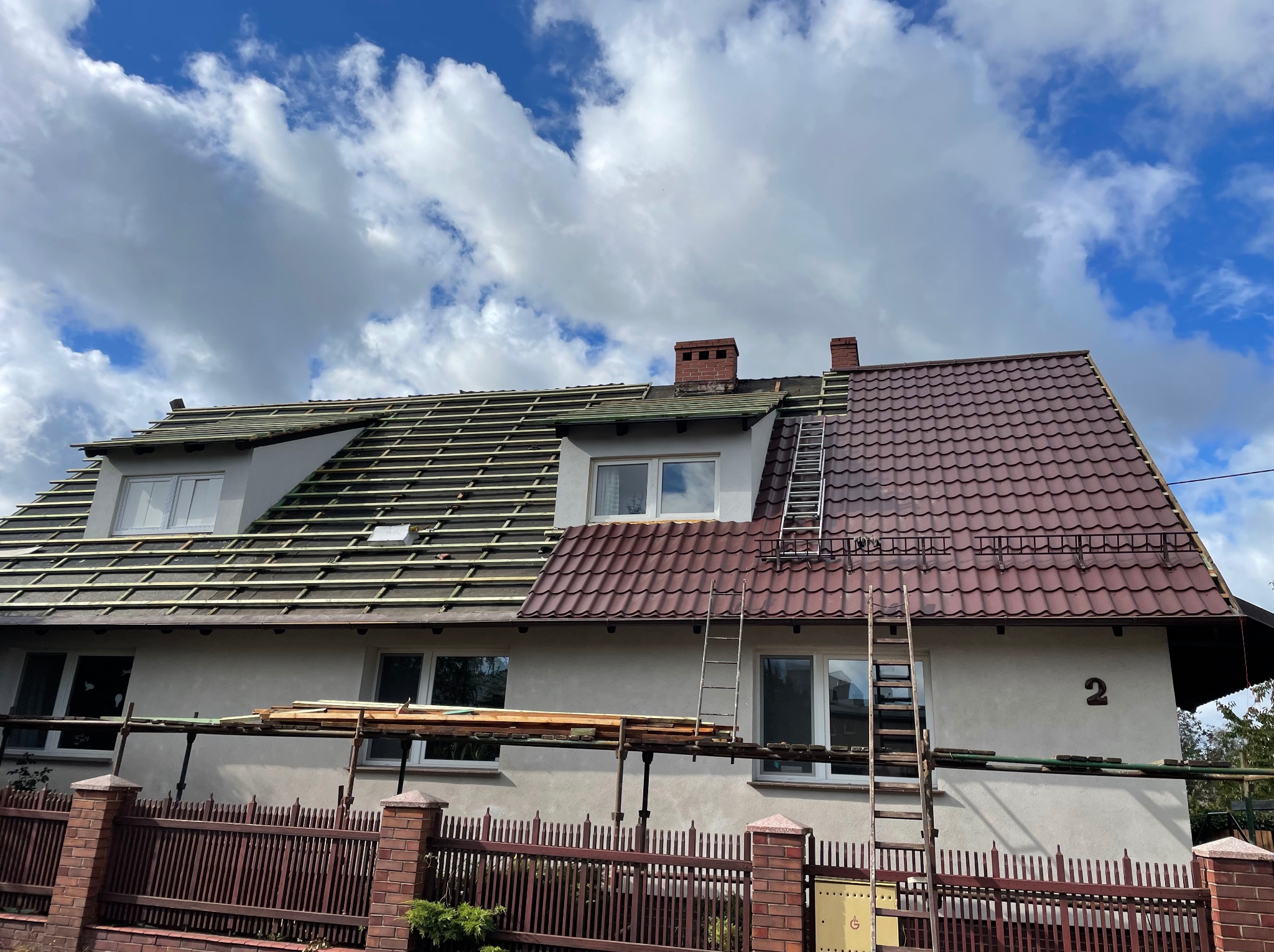 Dom jednorodzinny z częściowo zdjętym i częściowo nowym dachem z brązowej dachówki, rusztowanie, drabiny opierają się o ścianę, błękitne niebo z chmurami w tle.