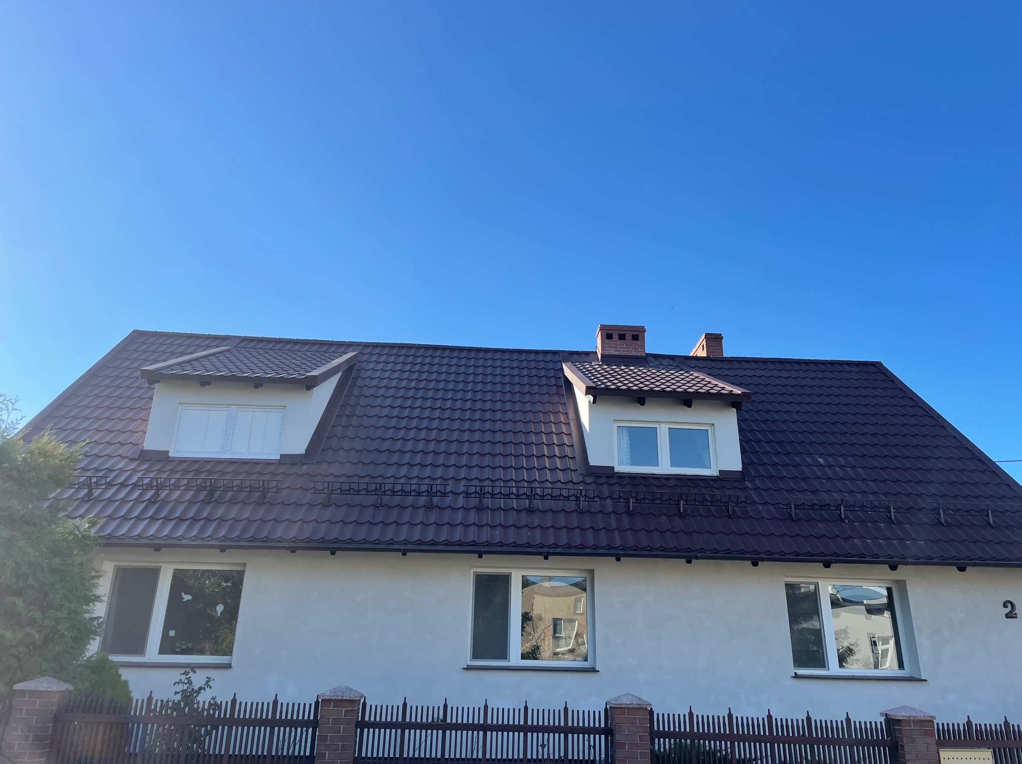 Dom jednorodzinny z brązowym dachem z dachówki, białymi oknami i kominem, widziany w słoneczny dzień z niskiej perspektywy.