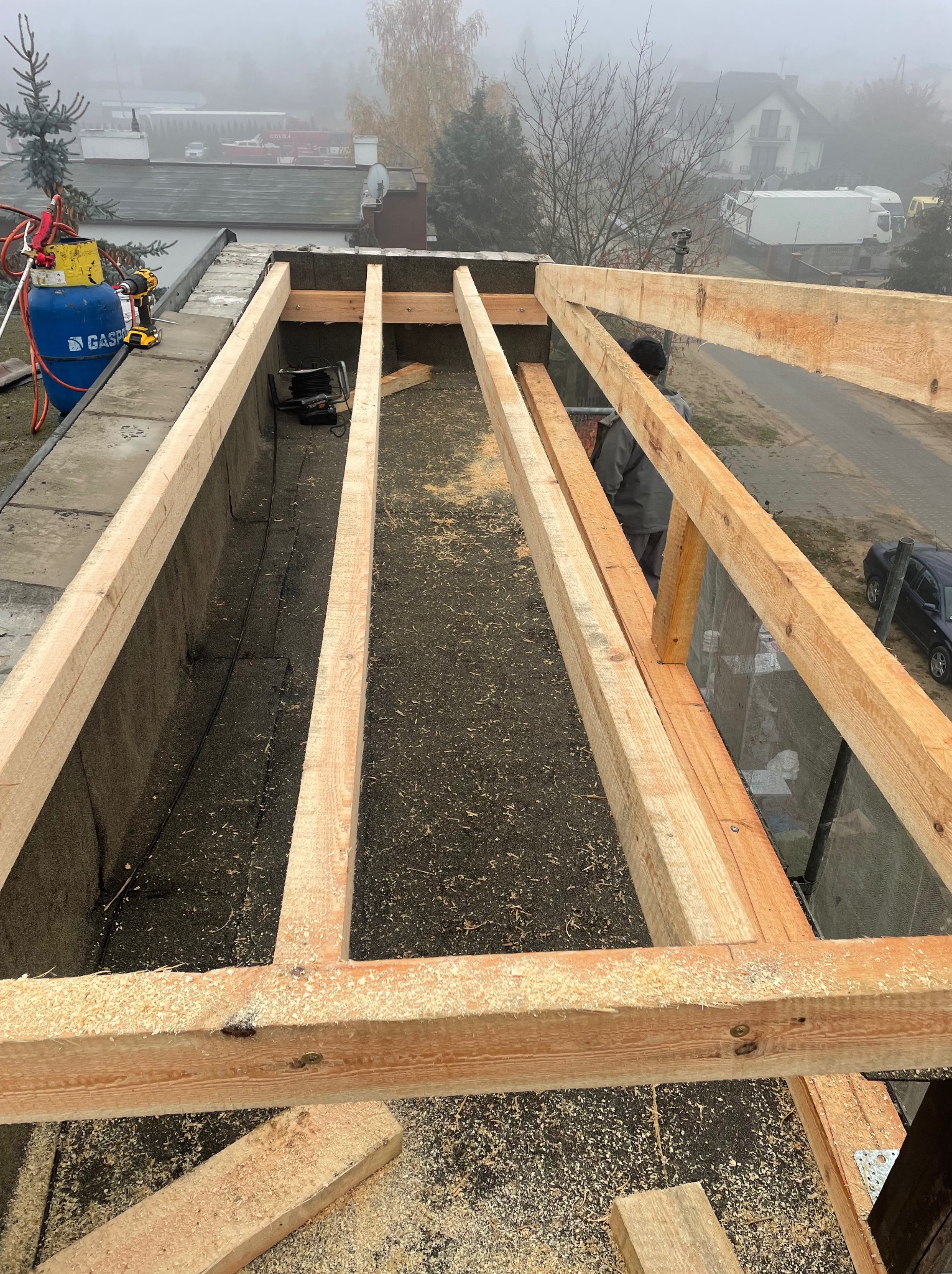 Konstrukcja drewniana dachu płaskiego w trakcie budowy, widoczne belki i deski, w tle budynki w porannej mgle, narzędzia i butla z gazem.