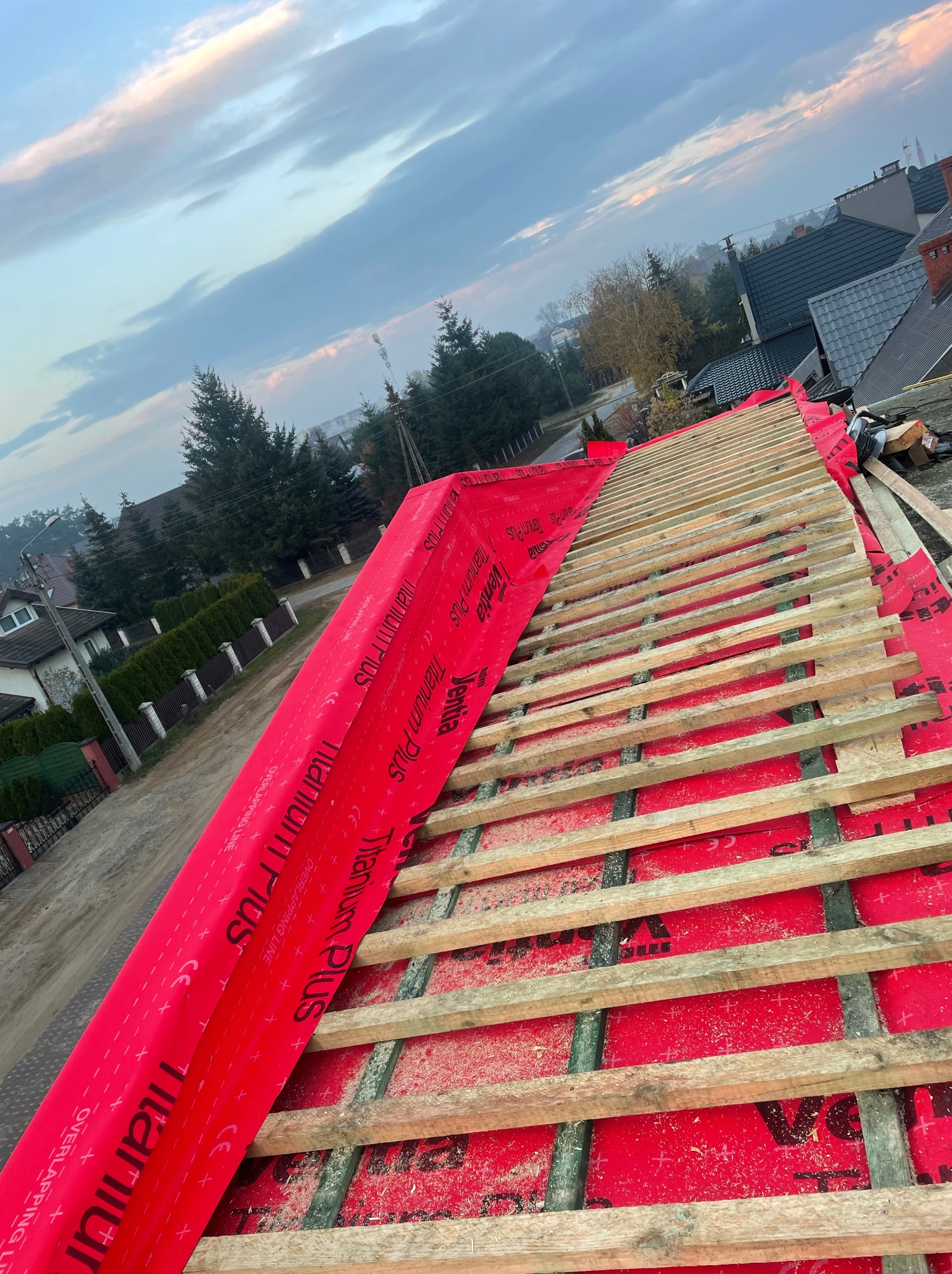 Konstrukcja dachu w trakcie budowy: czerwona membrana dachowa z nadrukiem 'Titanium Plus' rozciągnięta na krokwiach, widoczne deski łat, w tle zabudowa jednorodzinna i zachmurzone niebo.