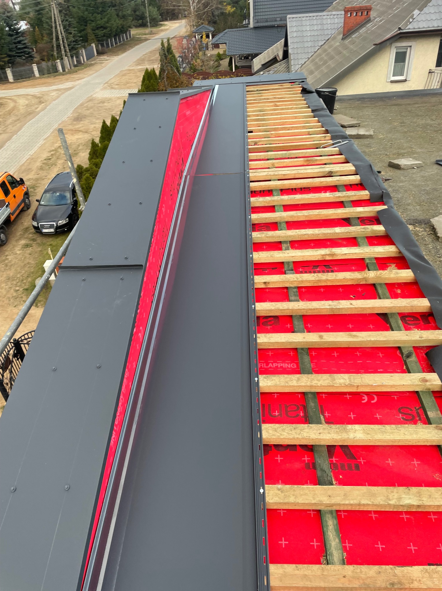 Montaż nowej, grafitowej blachy na dachu z widoczną czerwoną membraną i drewnianą konstrukcją łatowania. Widok z góry na dach w trakcie prac dekarskich.