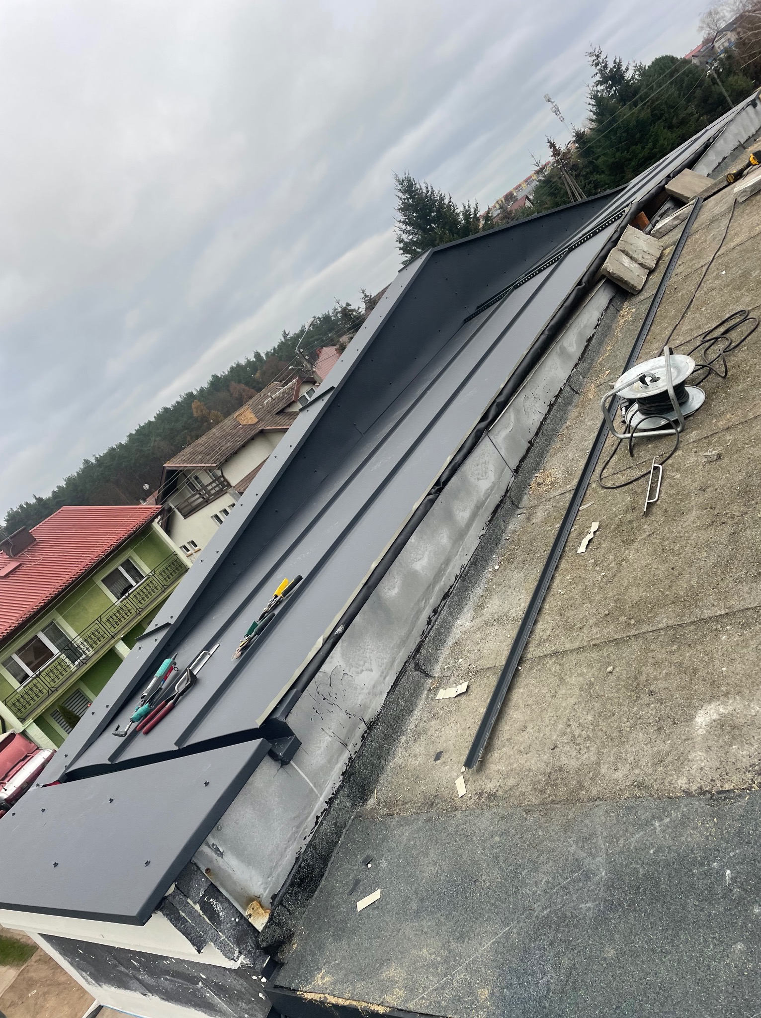 Montaż blachy na dachu płaskim, widoczne narzędzia dekarskie, zwój kabla, w tle zabudowa jednorodzinna i drzewa.