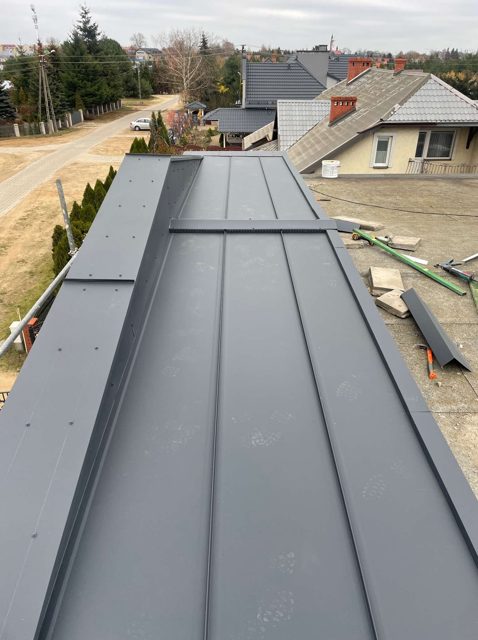 Nowoczesne, szare blaszane pokrycie dachowe z widocznymi śladami butów, narzędzia dekarskie i elementy konstrukcyjne na dachu w tle zabudowa jednorodzinna i drzewa.