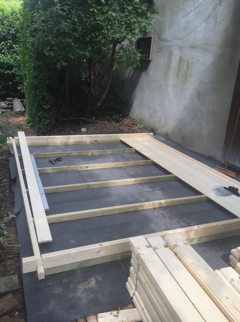 Konstrukcja drewnianego szkieletu pod podłogę, widoczna czarna membrana, narzędzia i stos desek na ziemi, obok ściana budynku i zielony krzew.