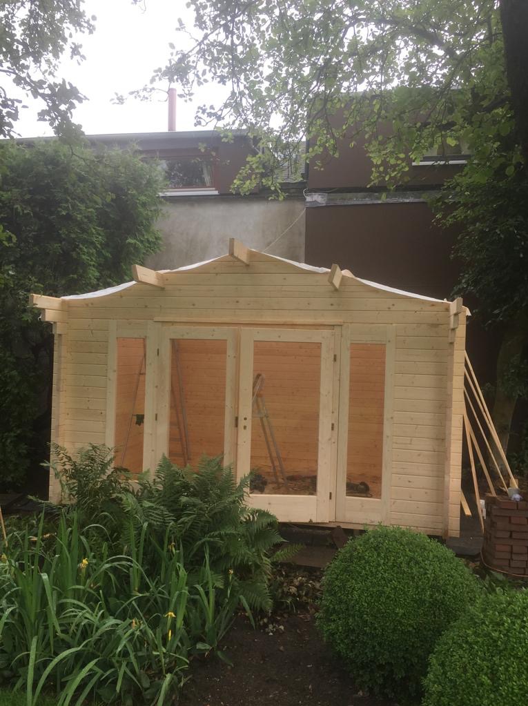 Montaż drewnianej konstrukcji altany ogrodowej z dwuskrzydłowymi drzwiami w Starogardzie Gdańskim, widoczne elementy konstrukcyjne dachu i cegły obok.