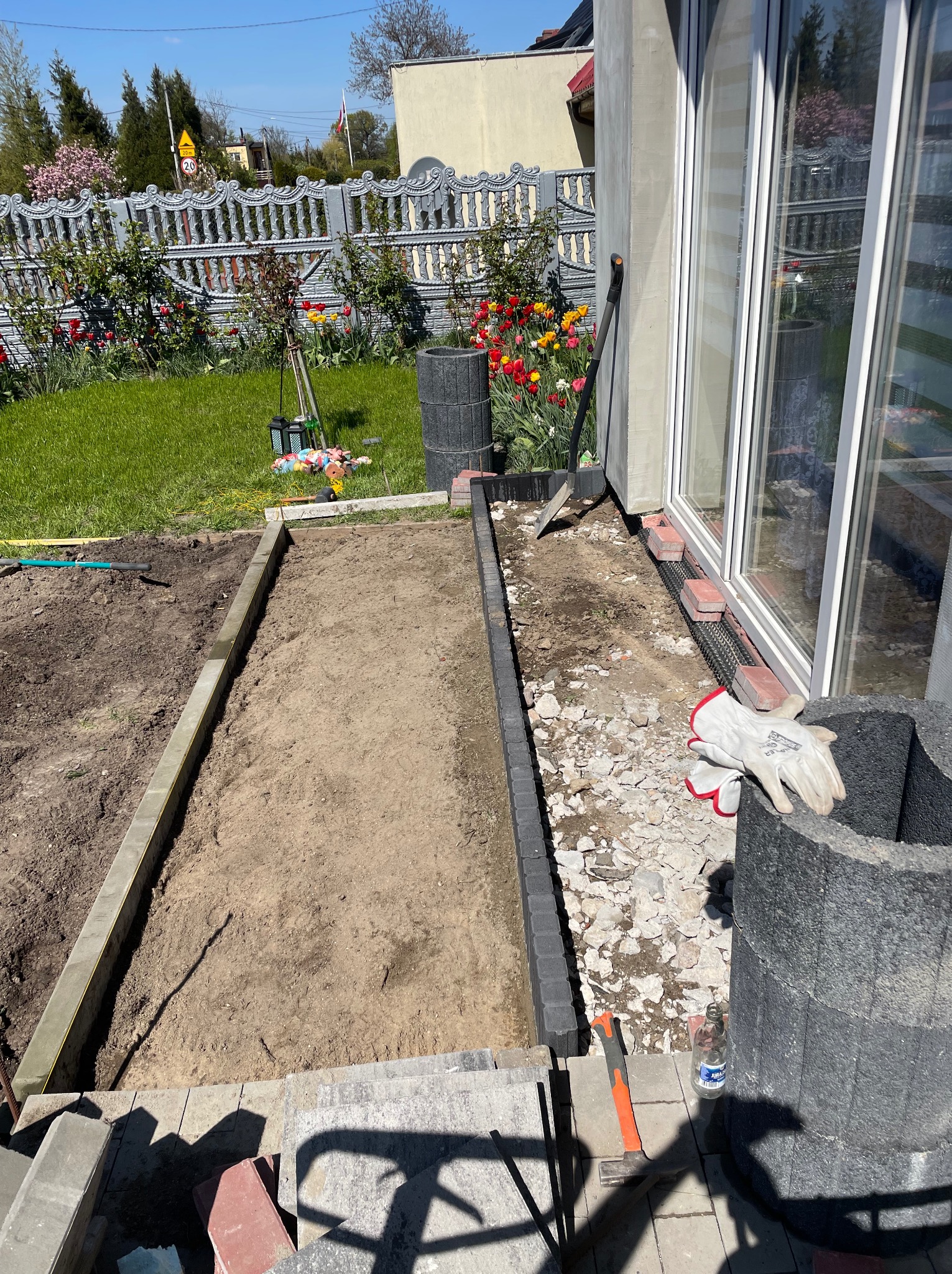 Przygotowanie podłoża pod taras: wykopane koryto z betonową obwódką, piasek, gruz, narzędzia i rękawice robocze przy ścianie budynku z dużymi oknami.