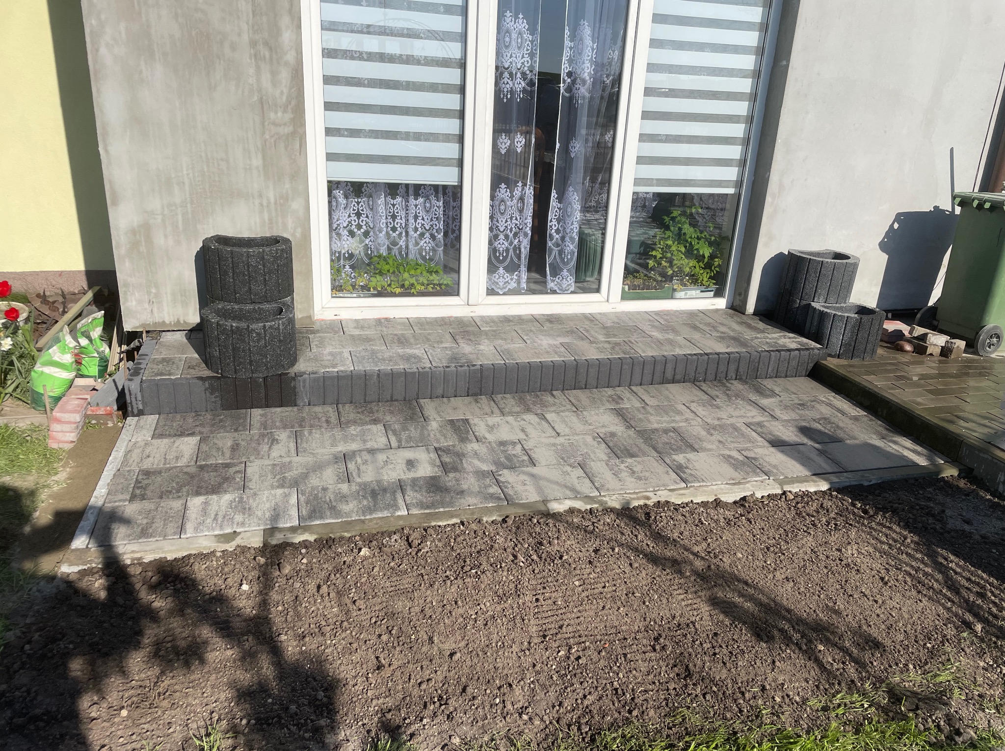 Ułożony z szarych kostek brukowych taras przed wejściem do domu z białymi drzwiami balkonowymi, obok ustawione czarne betonowe donice, widoczna świeżo przygotowana ziemia pod trawnik.