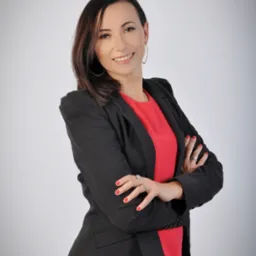 Uśmiechnięta kobieta w czarnej marynarce i czerwonej bluzce, z rękami skrzyżowanymi na piersiach, prezentująca ofertę ubezpieczeniową.