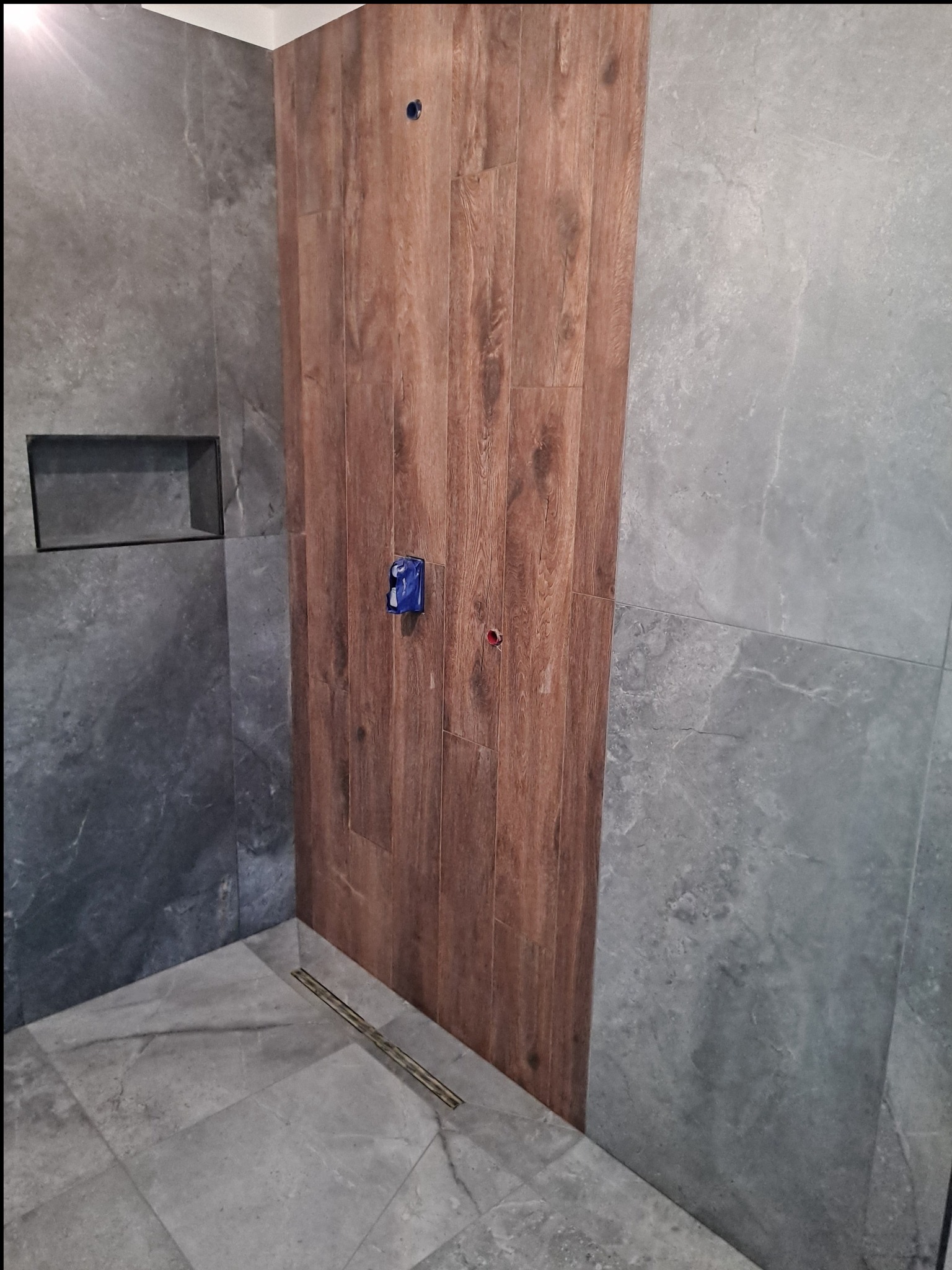 Fragment łazienki w trakcie wykańczania: ściana wyłożona płytkami imitującymi drewno sąsiaduje z szarą płytką imitującą kamień, widoczny odpływ liniowy w podłodze, zabezpieczone gniazdko elektryczne.