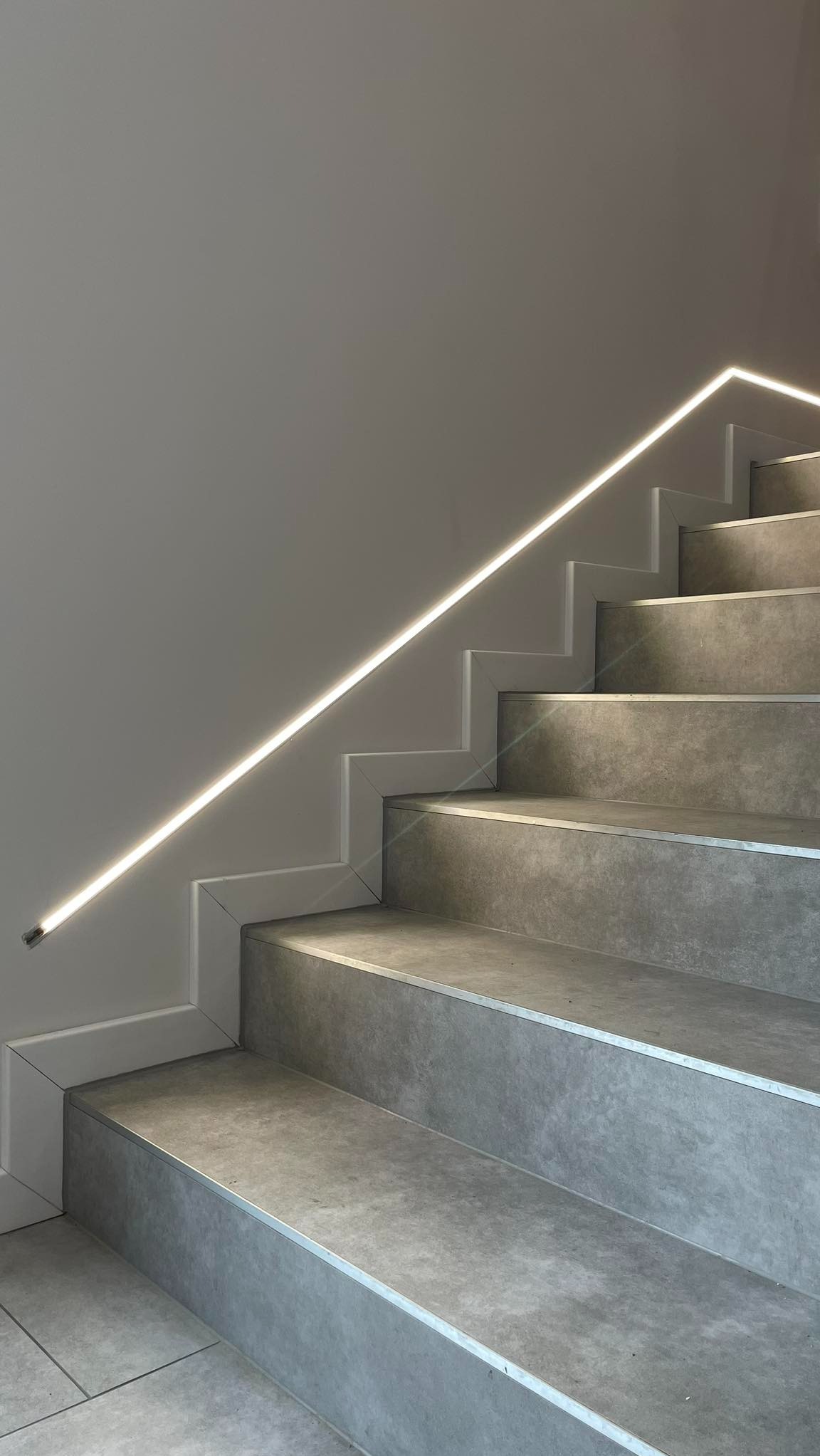 Nowoczesne schody z szarych płytek z podświetleniem LED wzdłuż ściany, minimalistyczny design.