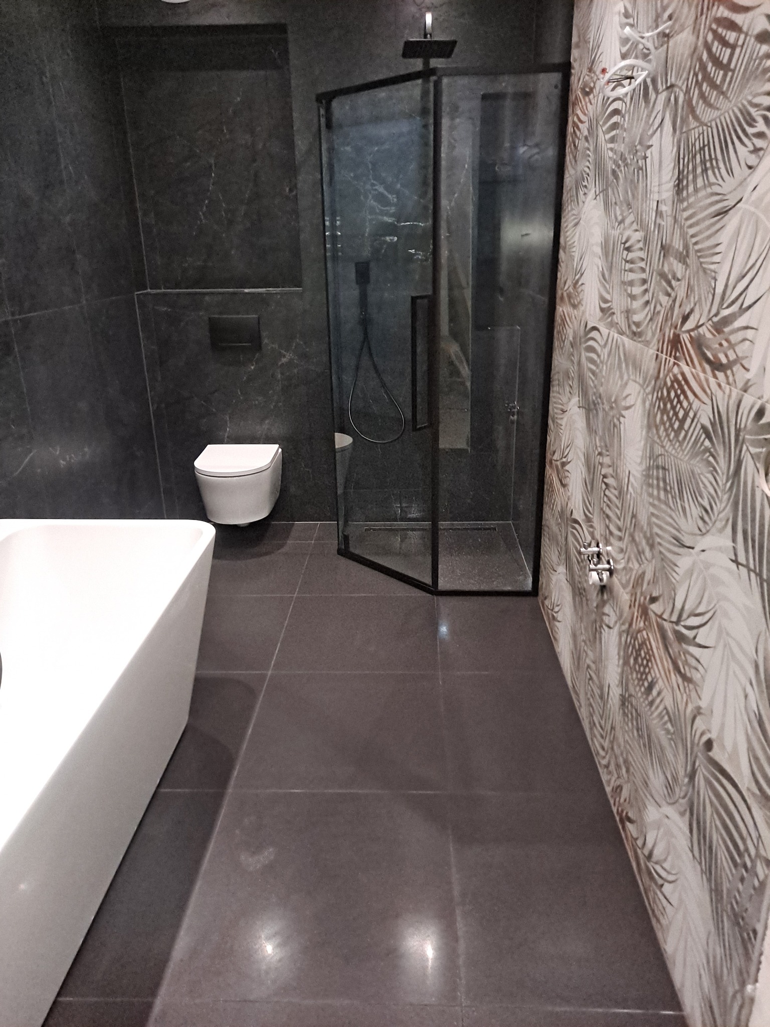 Nowoczesna łazienka z kabiną prysznicową typu walk-in z czarnymi profilami, wiszącą toaletą i wanną, wykończona ciemnymi płytkami imitującymi marmur oraz płytkami z motywem liści palmowych.