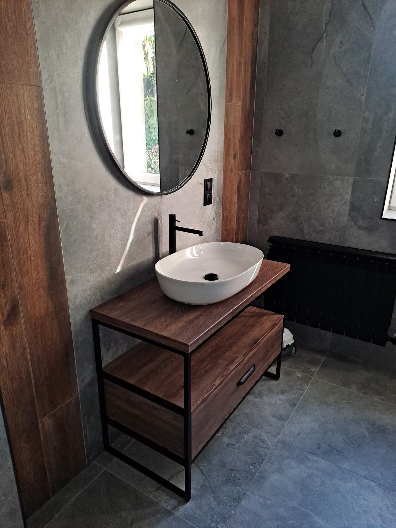 Nowoczesna łazienka z owalną umywalką nablatową na drewnianej szafce z czarną metalową ramą, okrągłe lustro w czarnej ramie, szare płytki na ścianach i podłodze, czarny grzejnik.