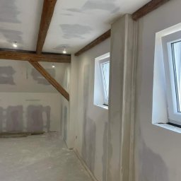 Instalacje elektryczne Zeus - Wnętrze w trakcie malowania: surowe drewniane belki stropowe, ściany pokryte gładzią szpachlową, nowe okna PCV, widoczne ślady po łataniu sufitu.