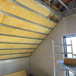 Instalacje elektryczne Zeus - Montaż izolacji z wełny mineralnej na poddaszu, widoczne metalowe profile i częściowo otynkowana ściana szczytowa, drabina rusztowaniowa w pomieszczeniu.