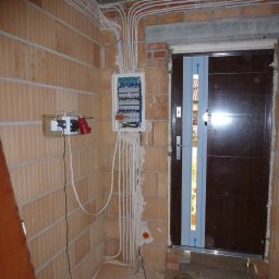 Instalacje elektryczne Zeus - Rozdzielnia elektryczna z licznymi przewodami w nowo budowanym pomieszczeniu z cegły, widoczne gniazda z wtyczkami i zabezpieczona folią nowa, brązowa stolarka drzwiowa.