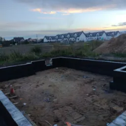 Fundamenty domu jednorodzinnego z hydroizolacją, widok na osiedle domów w tle, wieczorne niebo.