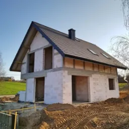 Surowy stan domu jednorodzinnego z białych bloczków, widoczna czarna dachówka i komin, teren wokół z wykopaną ziemią i śladami opon.