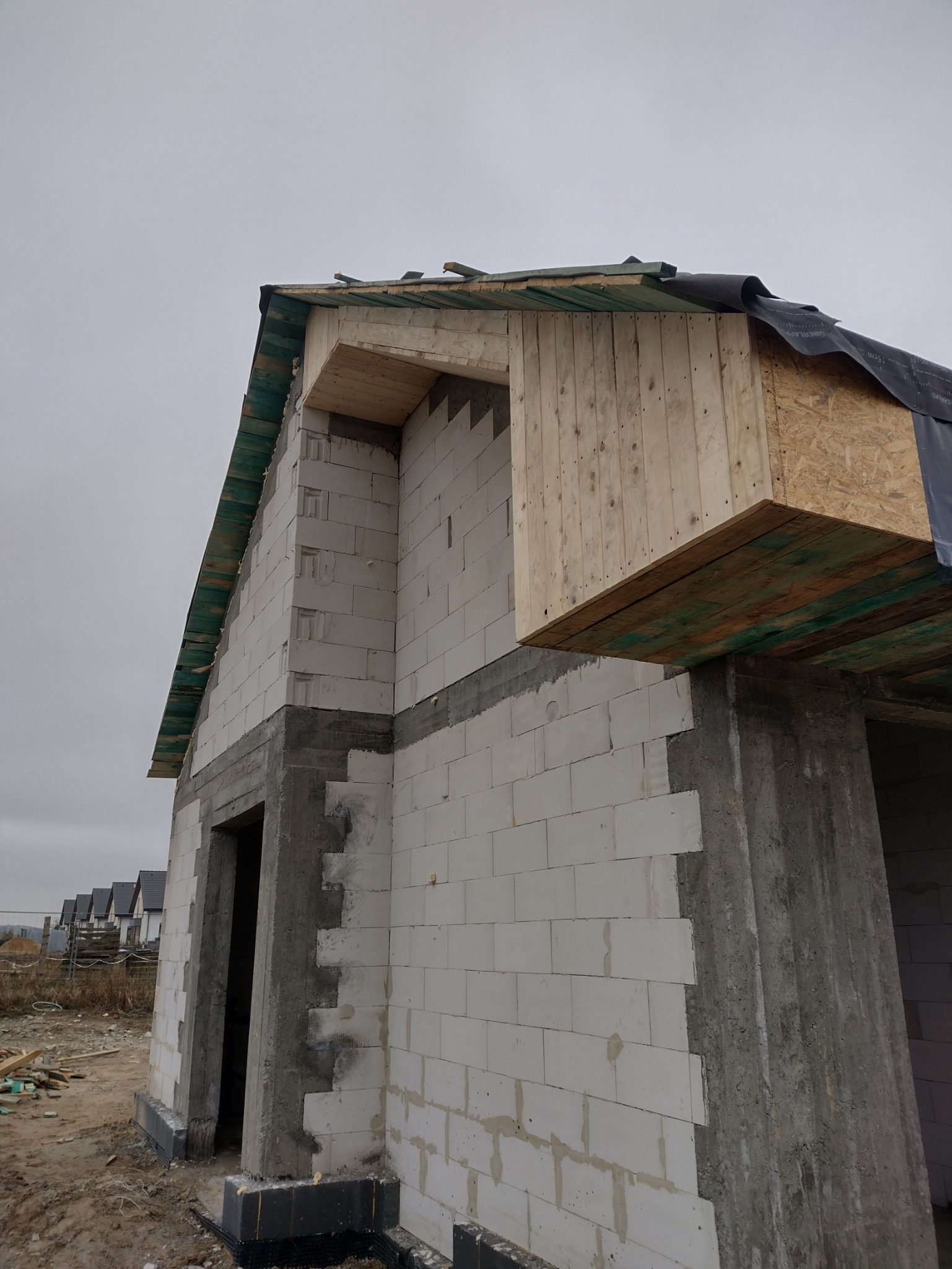 Surowy stan budowy domu jednorodzinnego: ściany z betonu komórkowego, drewniana konstrukcja dachu w trakcie montażu, widoczne elementy izolacji i hydroizolacji, w tle inne domy w zabudowie szeregowej.