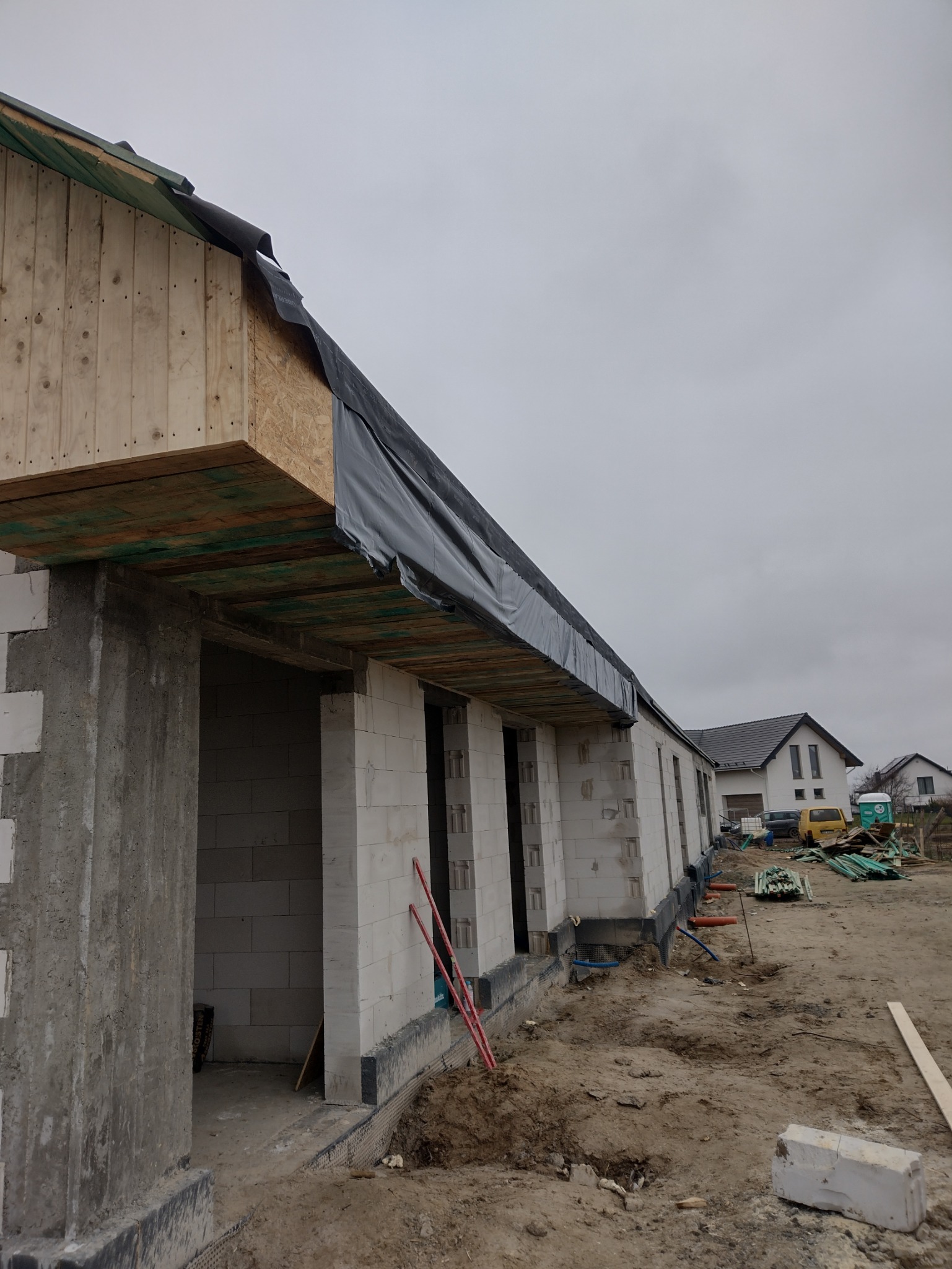 Surowy stan budynku jednorodzinnego z białych pustaków, z widoczną konstrukcją dachu z drewna i czarnej membrany, w tle inny dom i elementy budowlane.