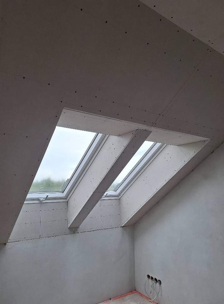 Wnętrze poddasza w trakcie wykańczania, widoczne okna dachowe w obudowie z płyt gipsowo-kartonowych, ściany przygotowane do malowania, wystające przewody elektryczne.