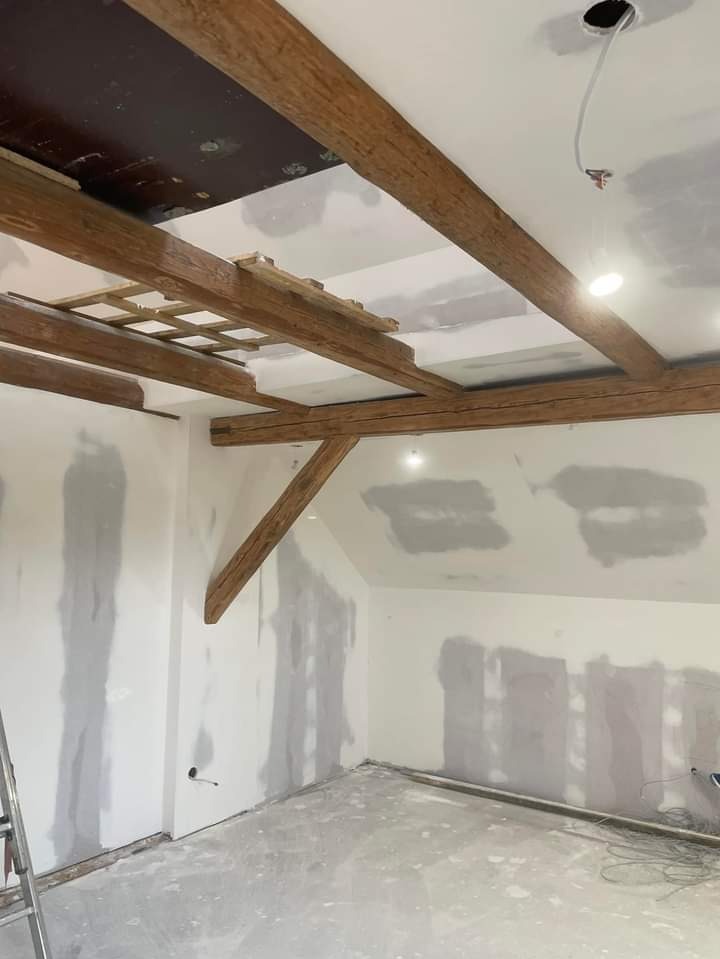 Pokój na poddaszu w trakcie prac wykończeniowych: widoczne drewniane belki stropowe, częściowo zamontowane płyty kartonowo-gipsowe, szpachlowane ściany i sufit, wystające przewody elektryczne...