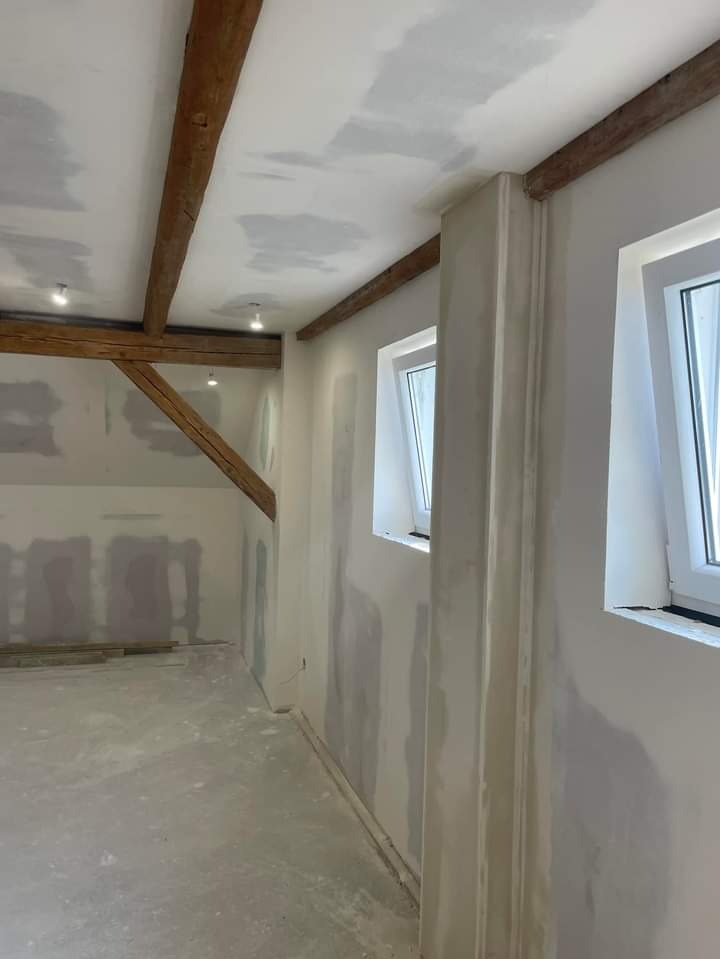 Wnętrze w trakcie malowania: surowe drewniane belki stropowe, ściany pokryte gładzią szpachlową, nowe okna PCV, widoczne ślady po łataniu sufitu.
