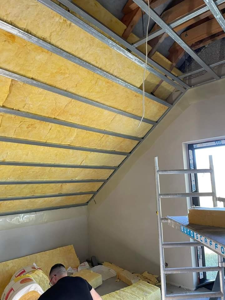 Montaż izolacji z wełny mineralnej na poddaszu, widoczne metalowe profile i częściowo otynkowana ściana szczytowa, drabina rusztowaniowa w pomieszczeniu.