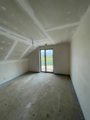 Pokój na poddaszu w trakcie wykańczania: zagruntowane ściany, płyty gipsowo-kartonowe na skosach, zabezpieczona folią podłoga, widok na pola za oknem.