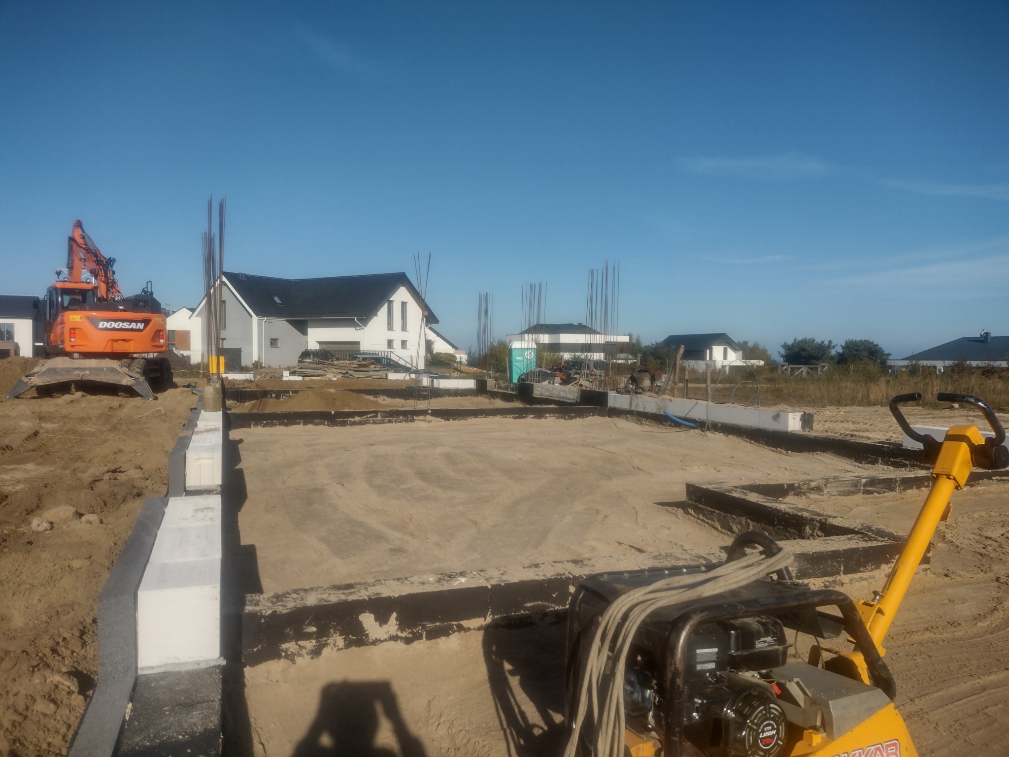 Przygotowanie fundamentów pod budynek jednorodzinny: wykop, szalunek z bloczków betonowych, zbrojenie pionowe, zagęszczarka i koparka Doosan w tle.