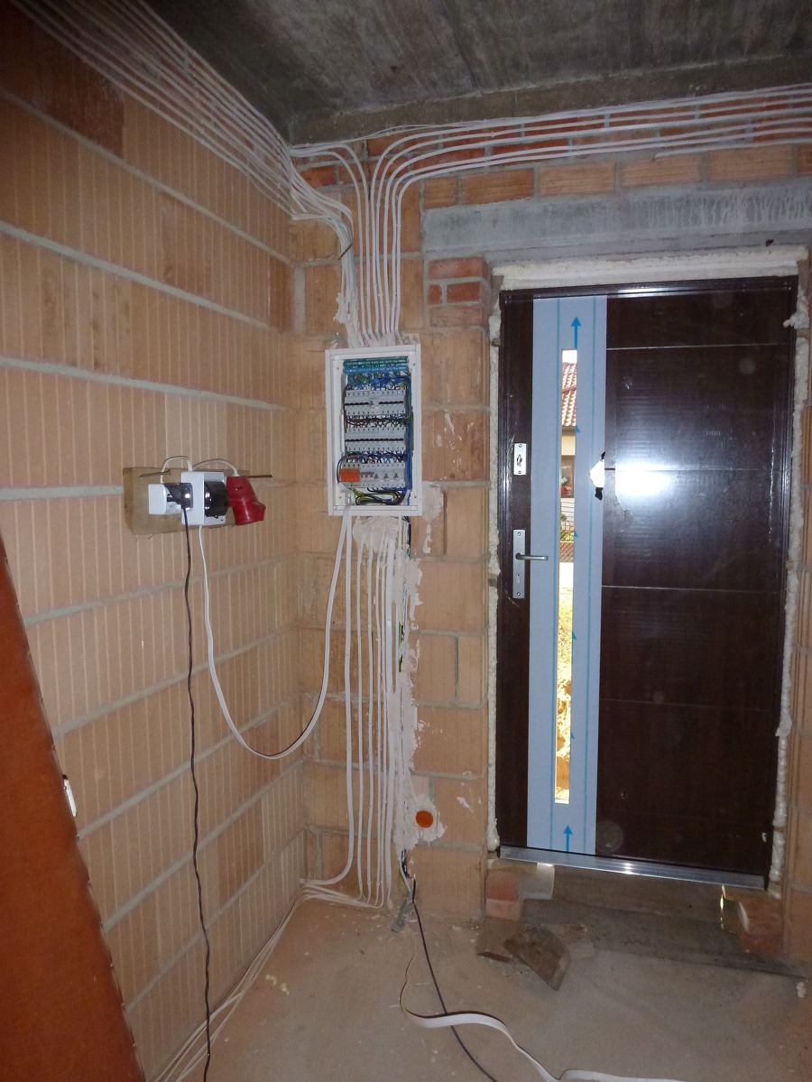 Rozdzielnia elektryczna z licznymi przewodami w nowo budowanym pomieszczeniu z cegły, widoczne gniazda z wtyczkami i zabezpieczona folią nowa, brązowa stolarka drzwiowa.