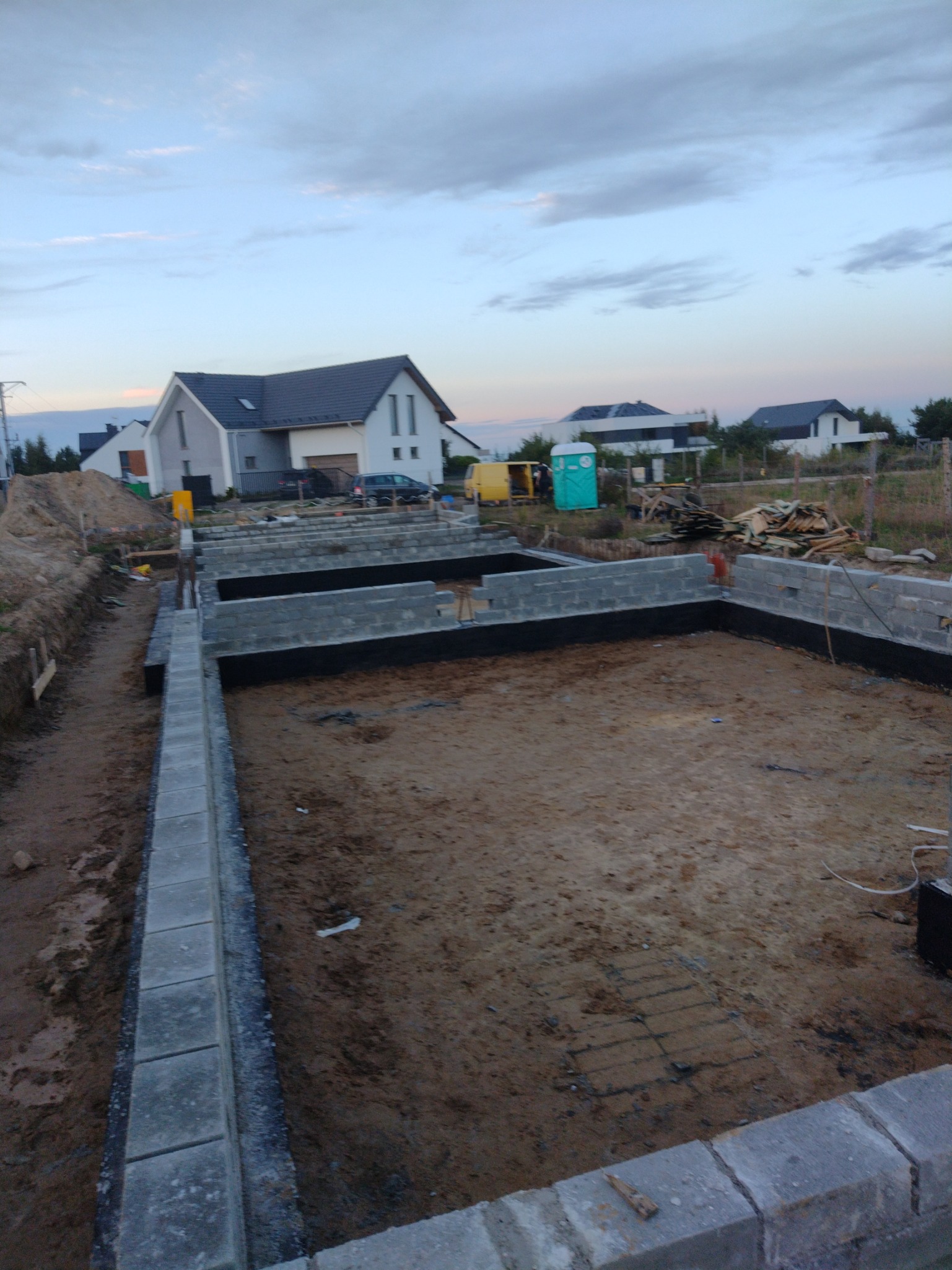 Fundamenty domu jednorodzinnego w trakcie budowy, widoczne bloczki betonowe, izolacja przeciwwilgociowa, zbrojenie w wykopie, w tle budynki mieszkalne i błękitne niebo.