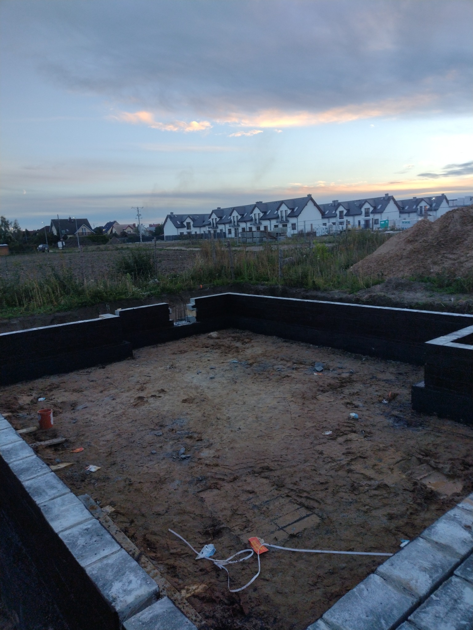 Fundamenty domu jednorodzinnego z hydroizolacją, widok na osiedle domów w tle, wieczorne niebo.
