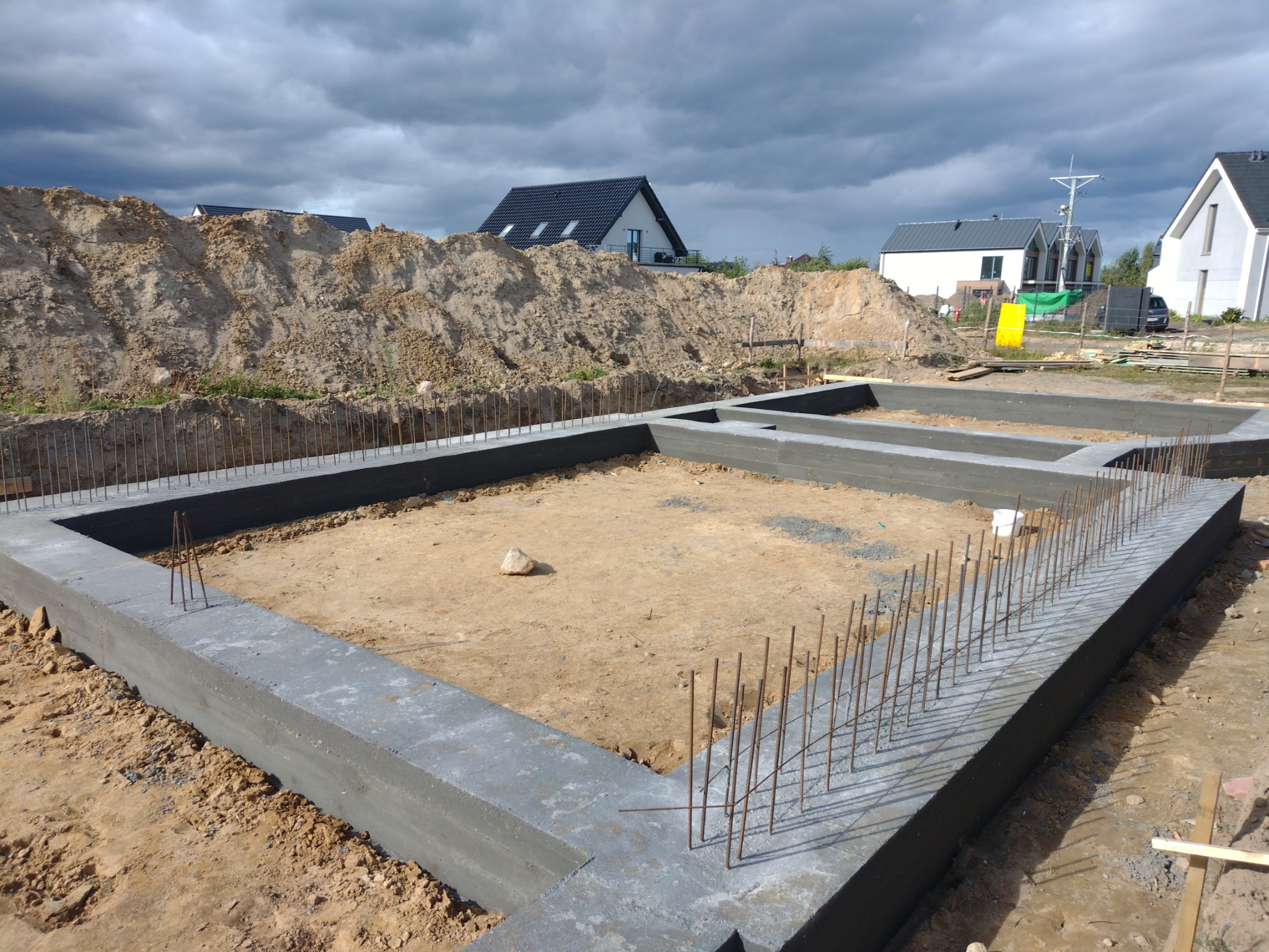 Wylane fundamenty domu jednorodzinnego z wystającymi prętami zbrojeniowymi, widok z góry na plac budowy z usypaną ziemią w tle i pochmurnym niebem.