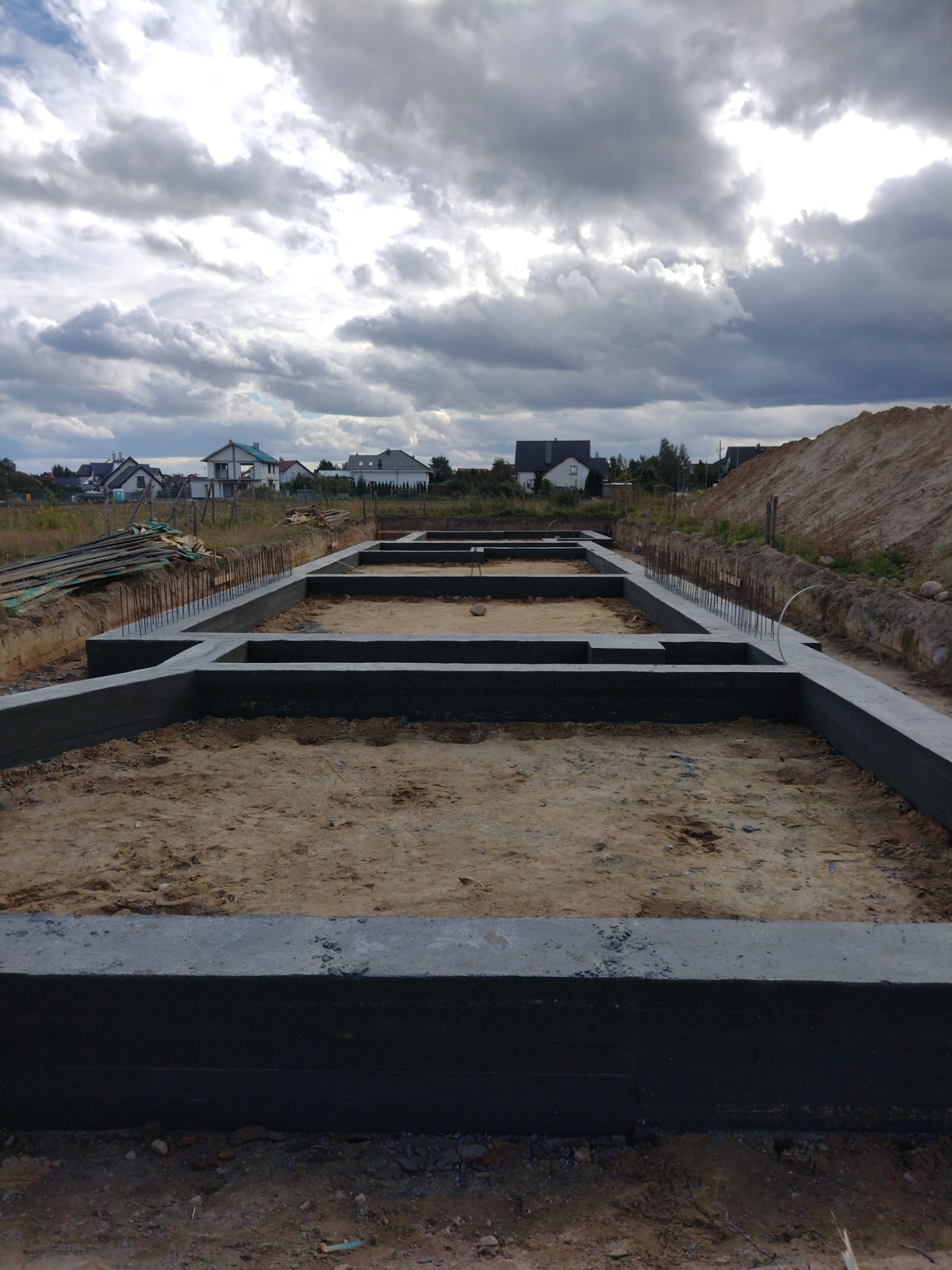 Wylane fundamenty domu jednorodzinnego z wystającymi prętami zbrojeniowymi, widok na plac budowy i pochmurne niebo.