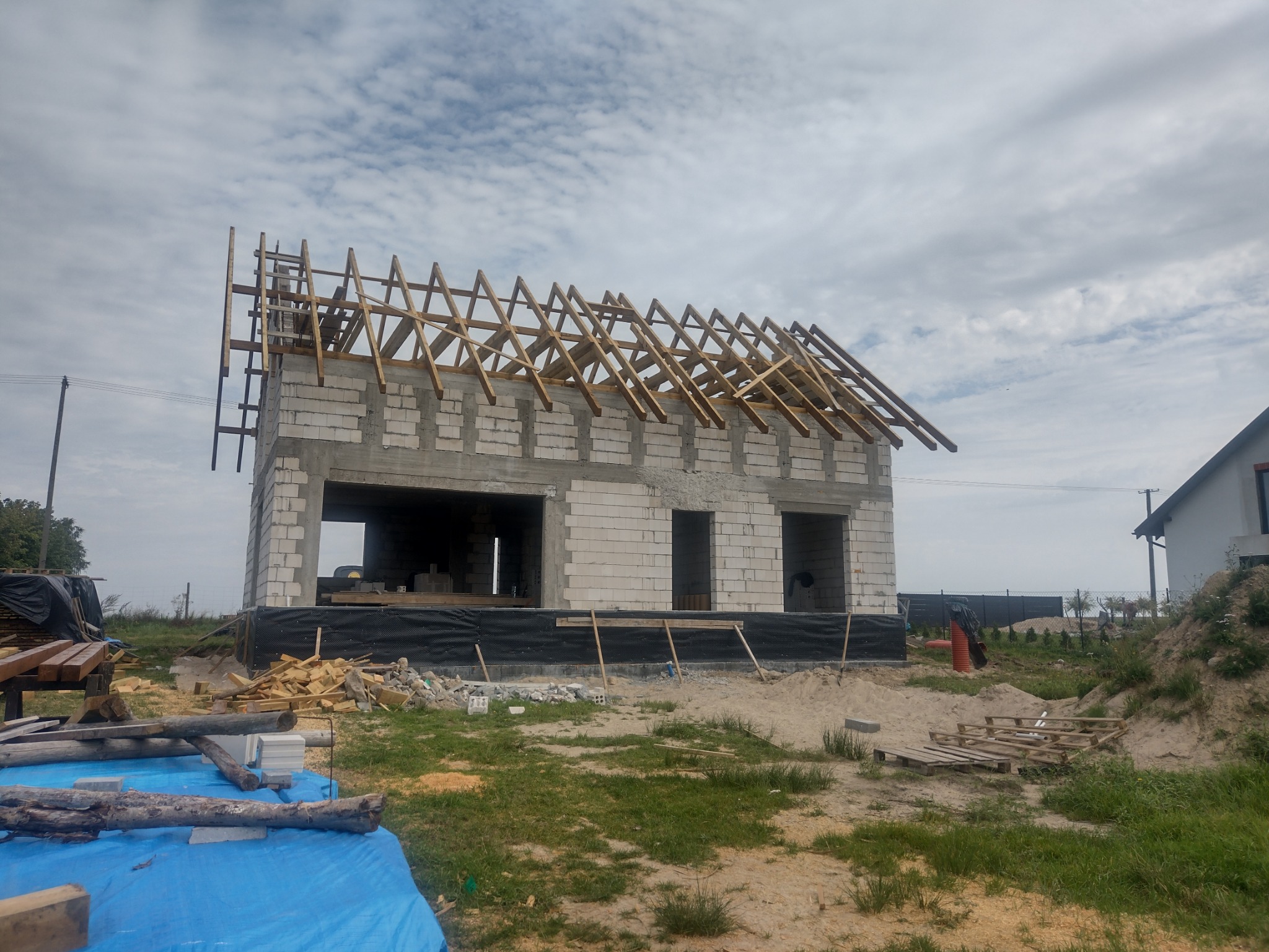 Surowy stan domu z białych bloków z drewnianą konstrukcją dachu w trakcie budowy, widoczne materiały budowlane i sąsiedni dom w tle.