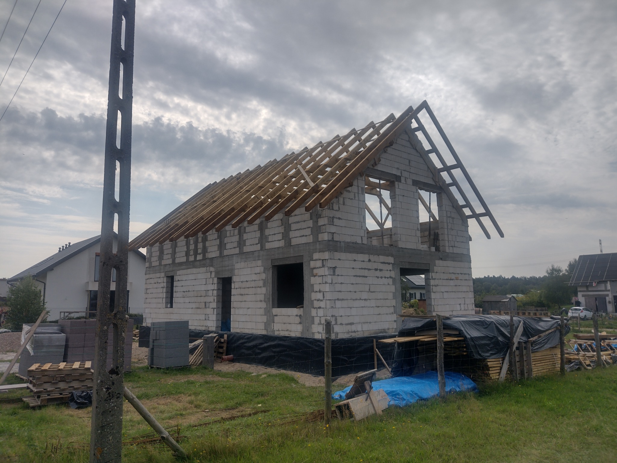 Surowy stan domu jednorodzinnego z pustaków, z widoczną konstrukcją dachu w trakcie budowy, na tle zachmurzonego nieba i sąsiednich budynków, z materiałami budowlanymi składowanymi na posesji.