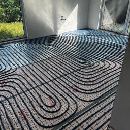Hydro-Bud Łukasz Bulanda - Instalacja ogrzewania podłogowego w nowym budynku, widoczne rury grzewcze ułożone na izolacji z siatką, jasne wnętrze z dużymi oknami.
