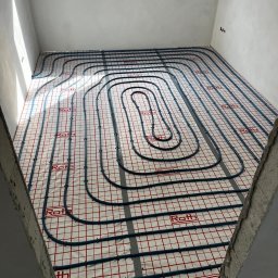 Hydro-Bud Łukasz Bulanda - Niebieskie rury ogrzewania podłogowego ułożone na czerwonej siatce w surowym stanie, widok z góry, w tle okno.