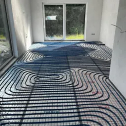 Rozłożona instalacja ogrzewania podłogowego z niebieskich rurek na folii z czerwoną siatką w surowym pomieszczeniu z dużymi oknami.