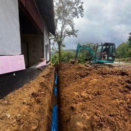 Hydro-Bud Łukasz Bulanda - Wykop pod rury, zabezpieczone niebieską folią, wykonany przy budynku z ociepleniem, z użyciem mini koparki.