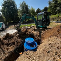 Hydro-Bud Łukasz Bulanda - Niebieski zbiornik na ścieki umieszczony w wykopie, obok stoi mała koparka, teren budowy otoczony zielenią.