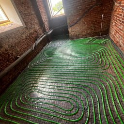 Hydro-Bud Łukasz Bulanda - Układanie zielonych rur ogrzewania podłogowego na folii z kratką w pomieszczeniu z ceglanymi ścianami i oknami dachowymi.